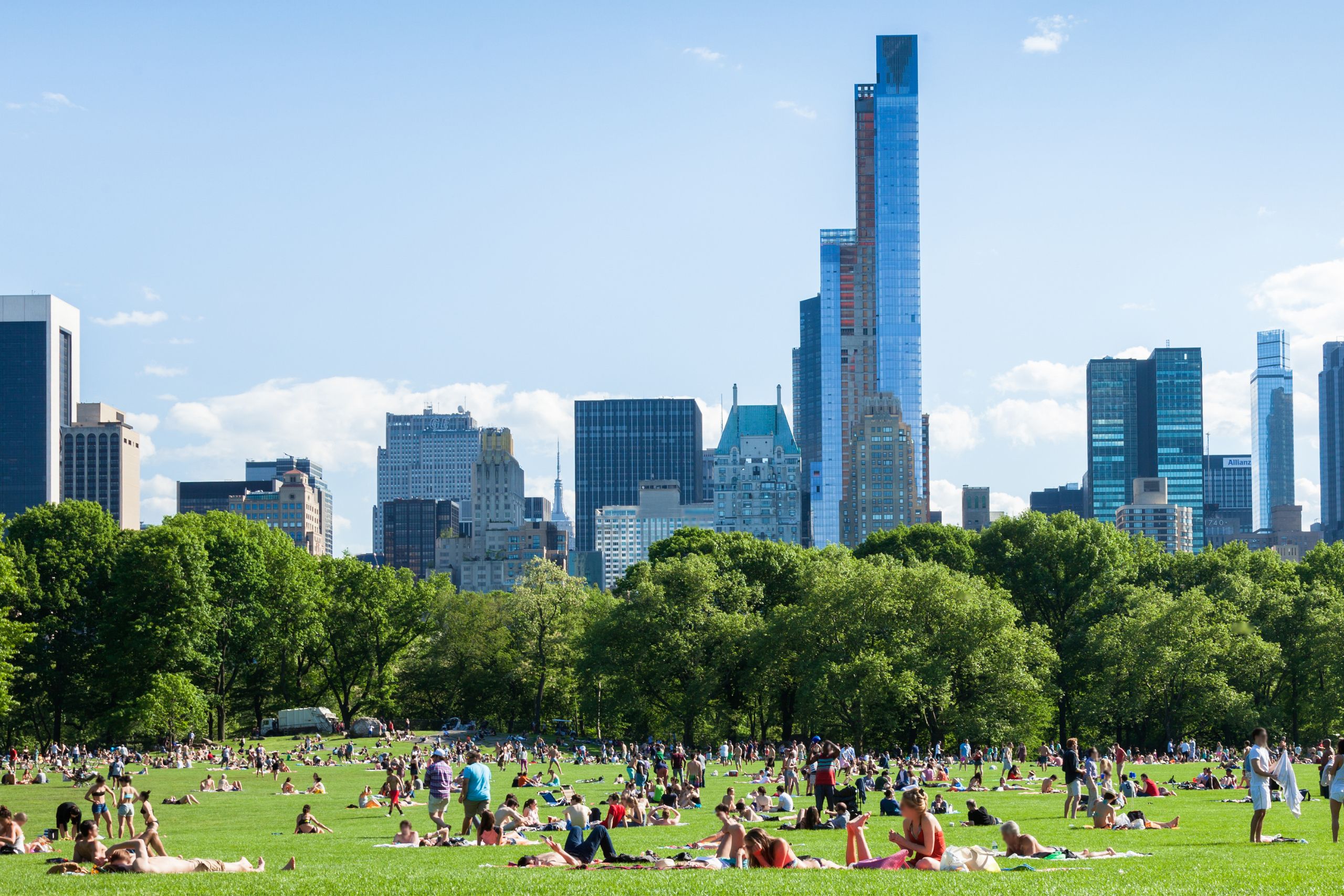 Crowded city park on a sunny day with a skyline of tall blue glass buildings behind the trees and people lounging on the grass.