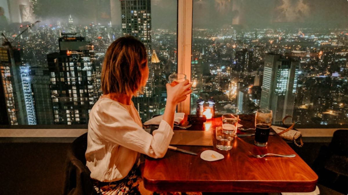 A woman enjoying rooftop dining in NYC with the Manhattan skyline glowing behind her at sunset